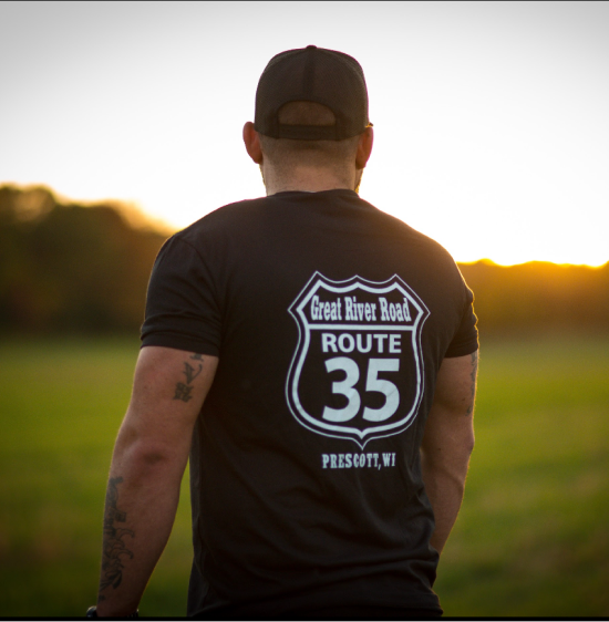 Man wearing a black t-shirt with 'Great River Road Route 35' design, standing in a field at sunset.