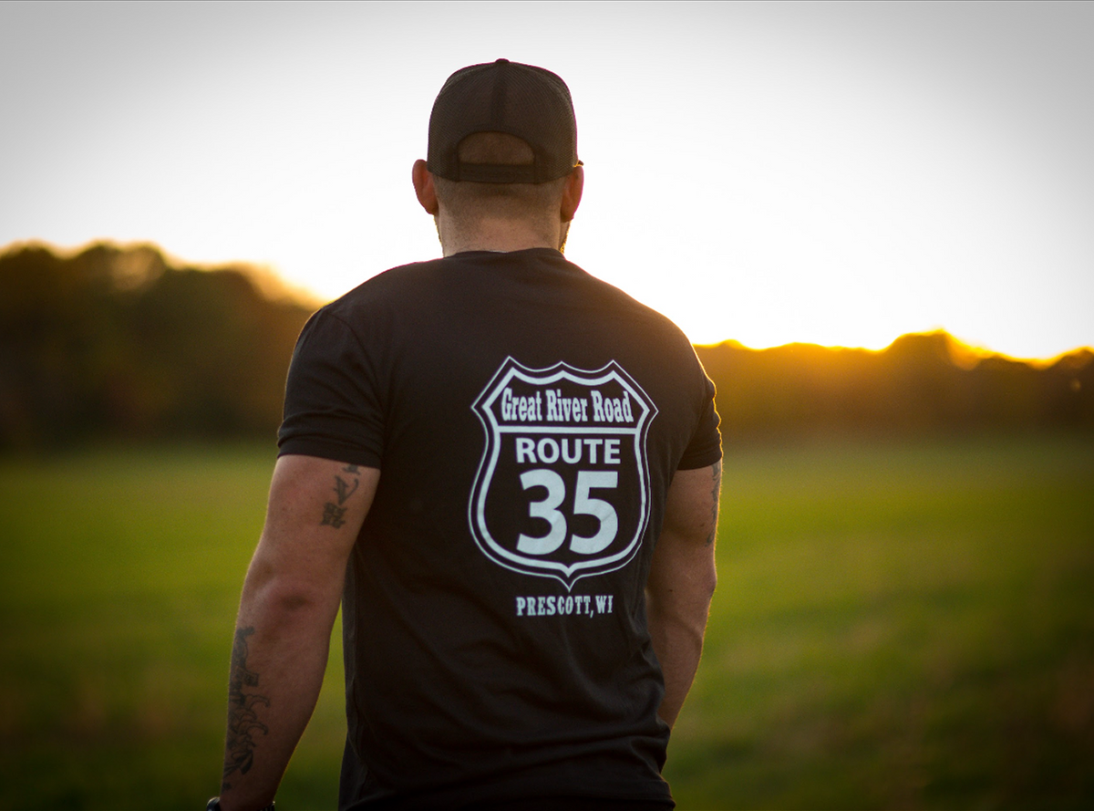 Person wearing a black t-shirt with 'Great River Road Route 35 Presque Isle, WI' design, standing in a field at sunset.
