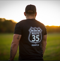 Man wearing a black t-shirt with 'Great River Road Route 35' design, standing in a field at sunset.